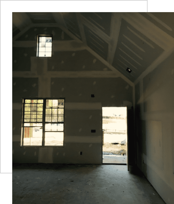 Interior of an unfinished room with drywall installed. Light enters through a door and two windows. Ceiling features exposed supports and partial plastering.